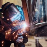 A skilled worker welding a rail segment in a metal fabrication workshop, with bright sparks flying from the welding point