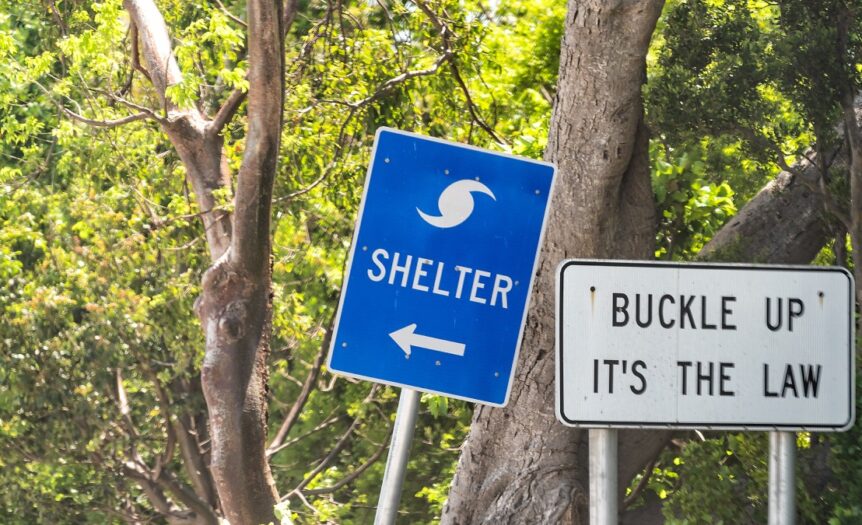 Two signs on a Florida road. One is blue and says "Shelter" with an arrow. The other is white and says "Buckle Up, It's the Law."