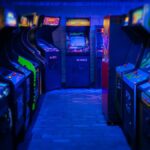 Two rows of retro arcade video games facing each other in a dark gaming room, with a blue light glowing from above.