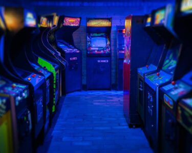 Two rows of retro arcade video games facing each other in a dark gaming room, with a blue light glowing from above.