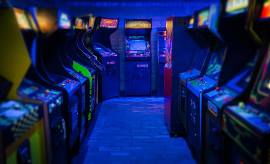 Two rows of retro arcade video games facing each other in a dark gaming room, with a blue light glowing from above.