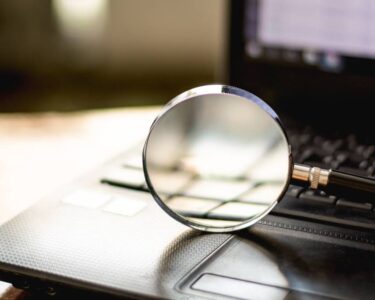 A black-handled magnifying glass rests on a black laptop's keyboard against a blurry meeting room background.