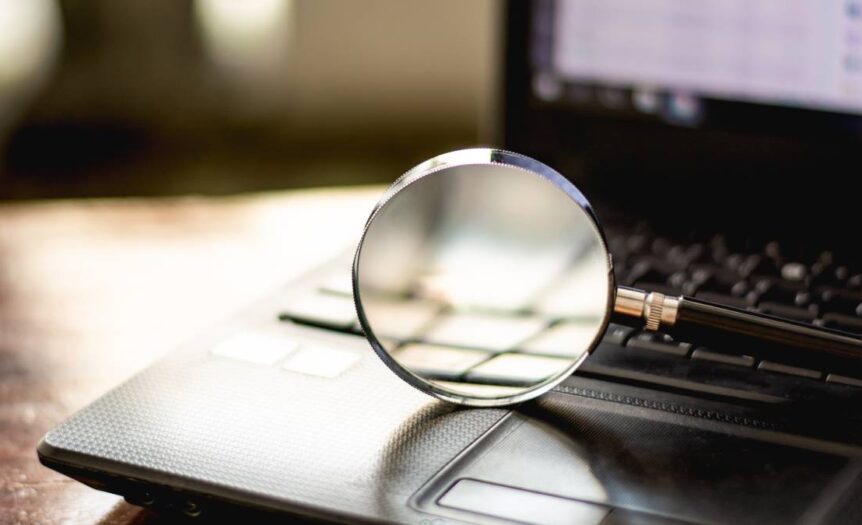 A black-handled magnifying glass rests on a black laptop's keyboard against a blurry meeting room background.