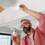 A man with a gray beard is changing the lightbulb in an overhead lamp. He is wearing glasses and a pink shirt.