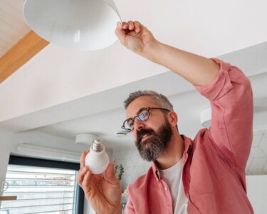 A man with a gray beard is changing the lightbulb in an overhead lamp. He is wearing glasses and a pink shirt.
