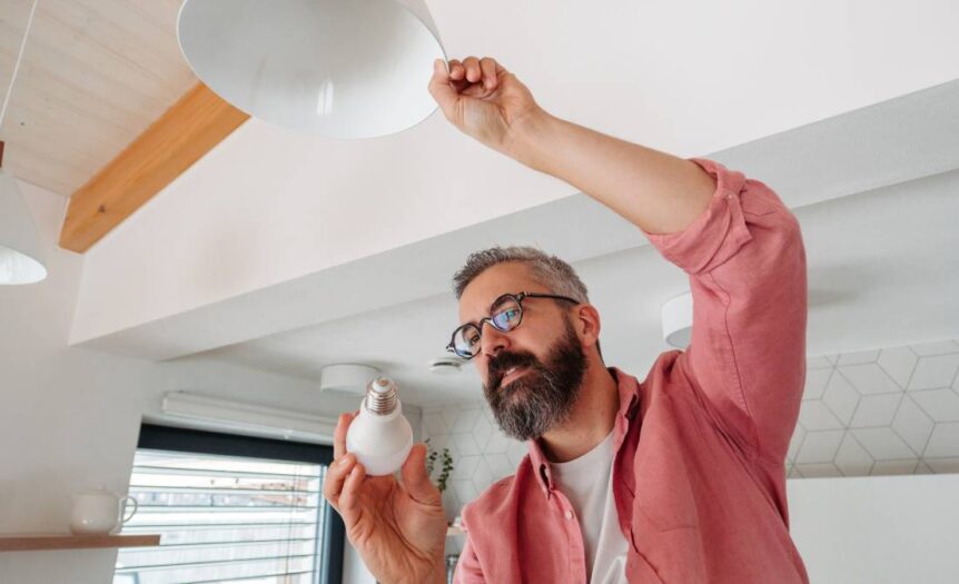 A man with a gray beard is changing the lightbulb in an overhead lamp. He is wearing glasses and a pink shirt.