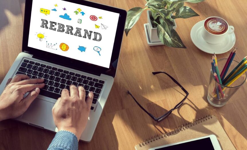 A laptop displaying a marketing rebrand on a table, accompanied by a plant, a cup of coffee, and a few pencils