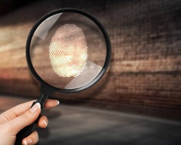 A woman's hand holding up a magnifying glass with a fingerprint on it in a dark, brick-walled alley.