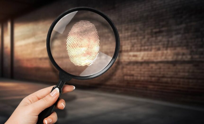 A woman's hand holding up a magnifying glass with a fingerprint on it in a dark, brick-walled alley.