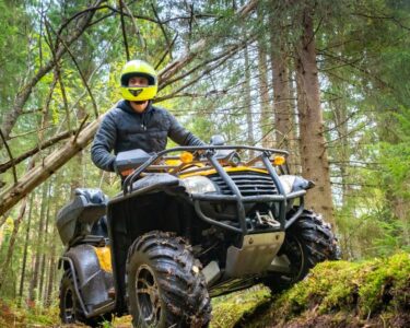 A man in a yellow helmet riding through the woods on a quad with large wheels covered in mud and leaves.