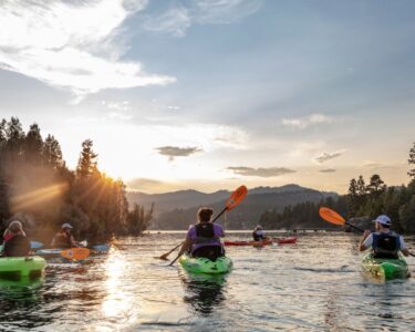 A family of five paddles their kayaks on a calm river as the sun sets in the background. Bluffs rise on the sides of the river.