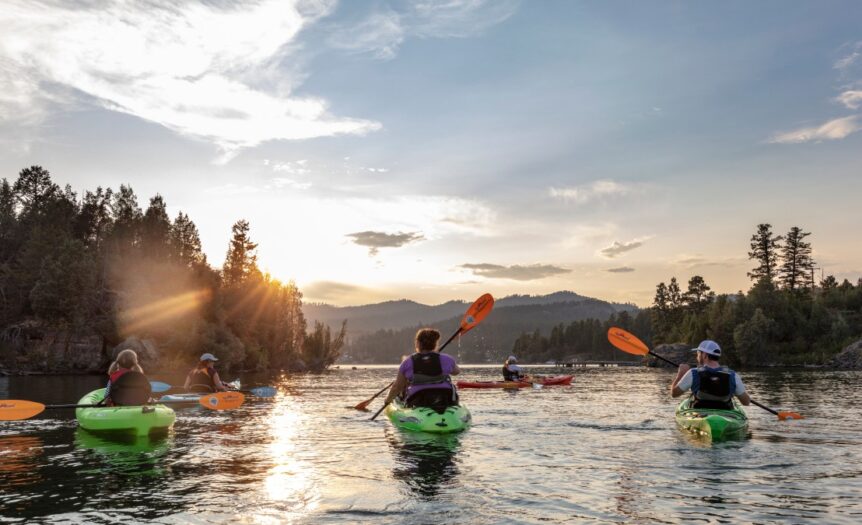 A family of five paddles their kayaks on a calm river as the sun sets in the background. Bluffs rise on the sides of the river.