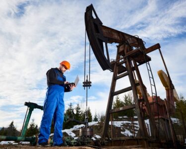 An oil worker in a hard hat and overalls using laptop to control or monitor the petroleum pump jack beside him.