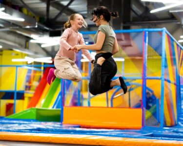 Two girls laughing and jumping in unison on a brightly colored trampoline inside a brightly lit playground park.