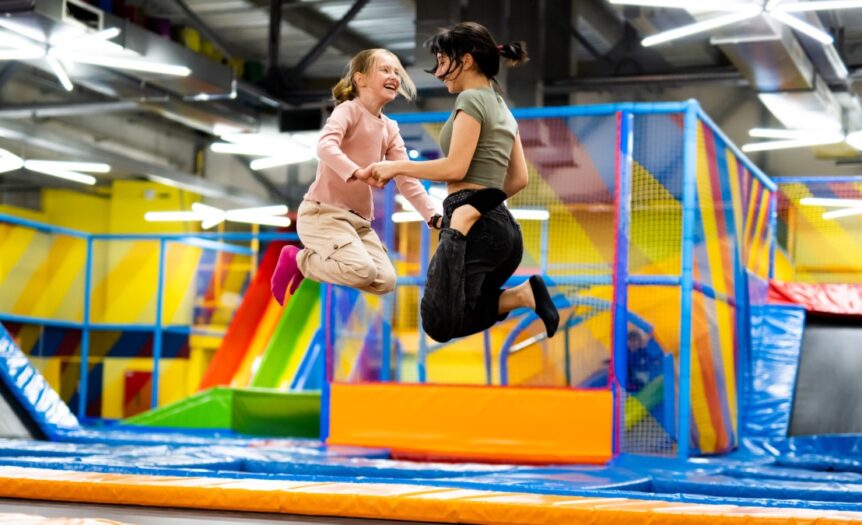 Two girls laughing and jumping in unison on a brightly colored trampoline inside a brightly lit playground park.