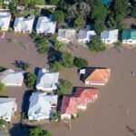 A bird’s eye view of a flooded suburban street. The houses' white, red, and green roofs stand out against the brown water.