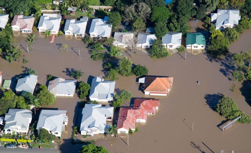 A bird’s eye view of a flooded suburban street. The houses' white, red, and green roofs stand out against the brown water.