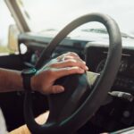 Someone sitting inside an older vehicle and resting their hand on the steering wheel’s horn button.