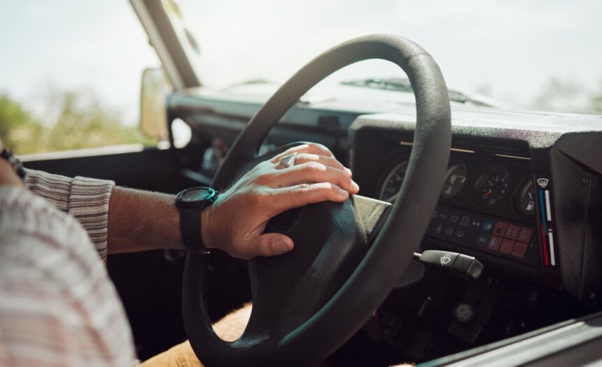 Someone sitting inside an older vehicle and resting their hand on the steering wheel’s horn button.