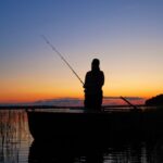 An angler holds a fishing rod while standing in a boat. The sun sets behind him in the background.