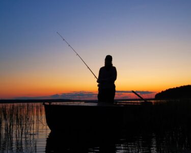 An angler holds a fishing rod while standing in a boat. The sun sets behind him in the background.