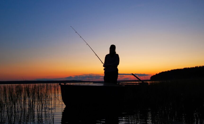 An angler holds a fishing rod while standing in a boat. The sun sets behind him in the background.