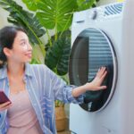 A woman is holding a smart phone in one hand while kneeling next to a smart washing machine. Her other hand is on the door.