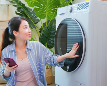 A woman is holding a smart phone in one hand while kneeling next to a smart washing machine. Her other hand is on the door.
