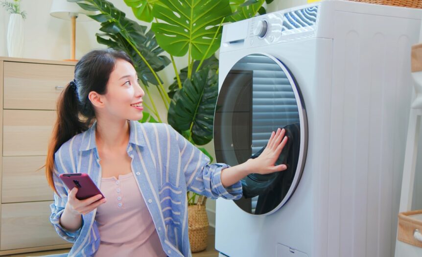 A woman is holding a smart phone in one hand while kneeling next to a smart washing machine. Her other hand is on the door.