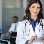 A doctor stands in a conference room and smiles while she holds a silver digital tablet in her hand.