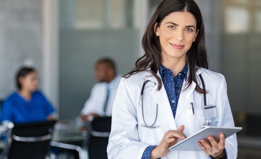 A doctor stands in a conference room and smiles while she holds a silver digital tablet in her hand.