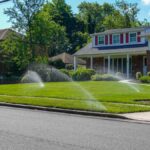 A lawn irrigation system in front of a suburban home spraying water from multiple nozzles across a lush, green front lawn.