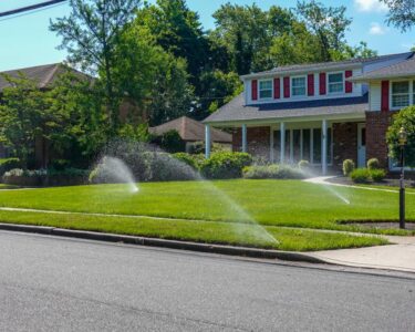 A lawn irrigation system in front of a suburban home spraying water from multiple nozzles across a lush, green front lawn.