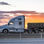 A semi-truck carrying cargo on its flatbed drives on the highway. The sun is setting in the background.