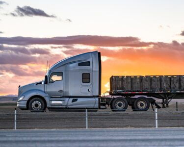 A semi-truck carrying cargo on its flatbed drives on the highway. The sun is setting in the background.