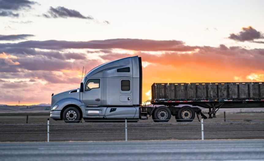 A semi-truck carrying cargo on its flatbed drives on the highway. The sun is setting in the background.