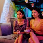 Two girls sit on a couch watching a movie together. The girl on the right has a bowl of food in her hand.