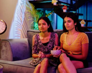 Two girls sit on a couch watching a movie together. The girl on the right has a bowl of food in her hand.