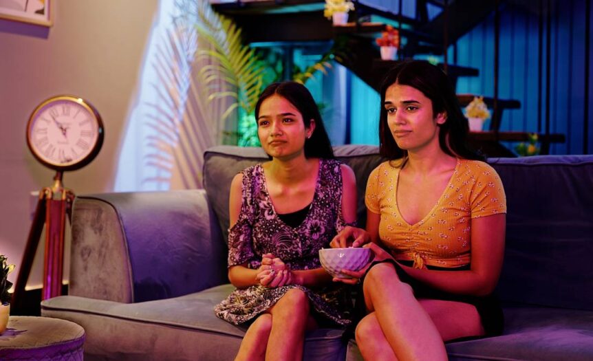 Two girls sit on a couch watching a movie together. The girl on the right has a bowl of food in her hand.