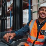 A young forklift driver operating a vehicle in a busy warehouse, smiling and looking confidently at the camera.