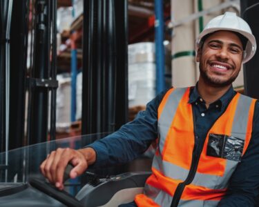 A young forklift driver operating a vehicle in a busy warehouse, smiling and looking confidently at the camera.