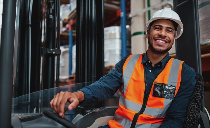 A young forklift driver operating a vehicle in a busy warehouse, smiling and looking confidently at the camera.