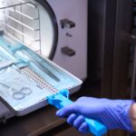 A hand wearing a blue glove inserts medical instruments into an autoclave to be sterilized.