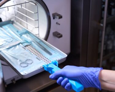 A hand wearing a blue glove inserts medical instruments into an autoclave to be sterilized.