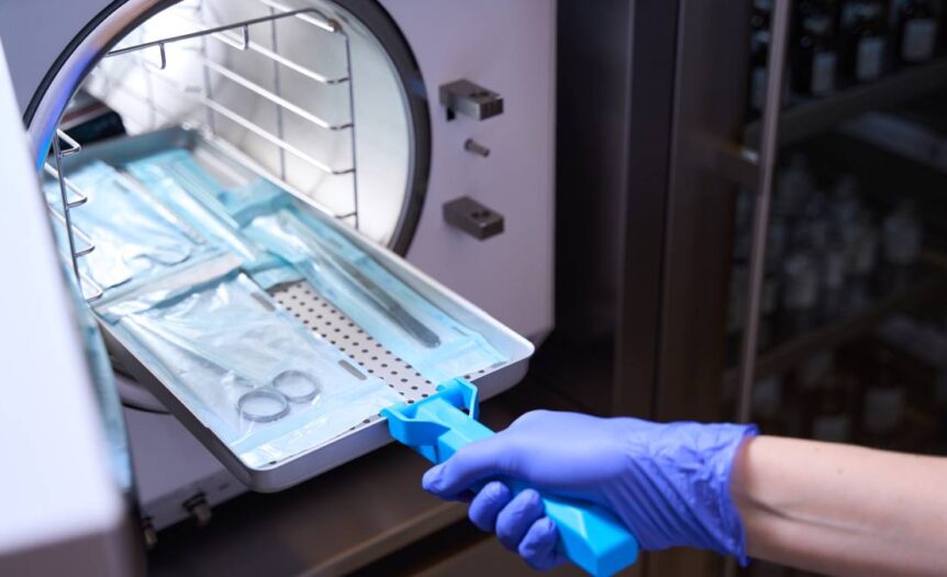 A hand wearing a blue glove inserts medical instruments into an autoclave to be sterilized.