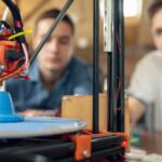 Two men sitting near a 3D printing machine set up on a brown table. The machine is printing a blue plastic figurine.