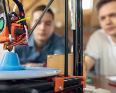 Two men sitting near a 3D printing machine set up on a brown table. The machine is printing a blue plastic figurine.