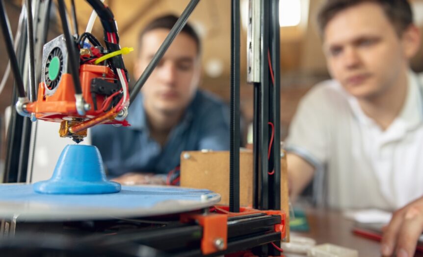 Two men sitting near a 3D printing machine set up on a brown table. The machine is printing a blue plastic figurine.