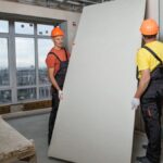 Two men wearing safety helmets and protective gear install drywall in a building under construction.