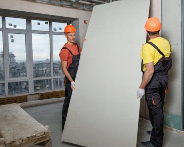 Two men wearing safety helmets and protective gear install drywall in a building under construction.